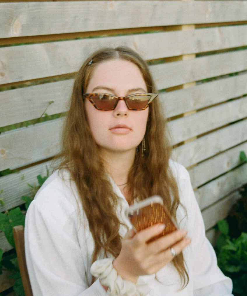 Portrait photograph of woman wearing sunglasses looking in camera whilst holding phone 