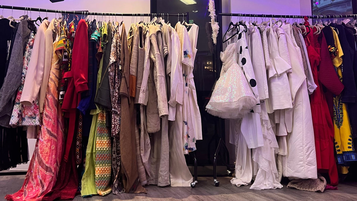 A clothing rack filled with assorted garments in neutral and bright colors inside a retail space. A large wall sign reads 'the SHACK', and another poster on the wall says 'ONE WEAR FREEDOM'.